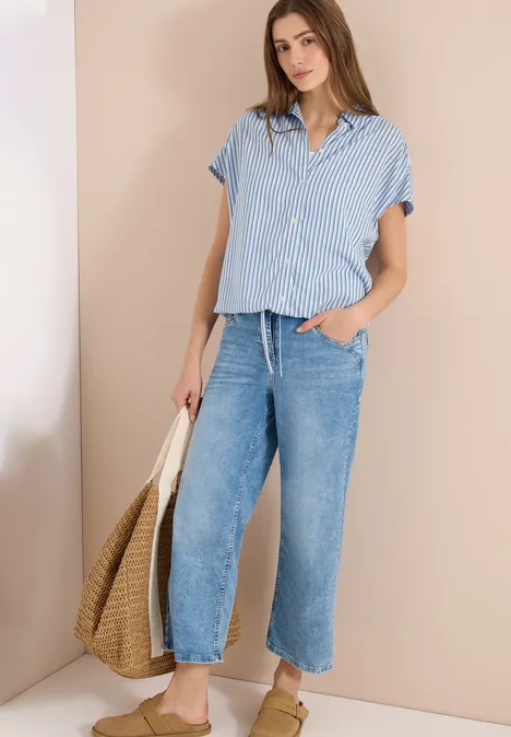 Vrouw in blauw-wit gestreept overhemd, wijde jeans en strozak voor beige achtergrond.
