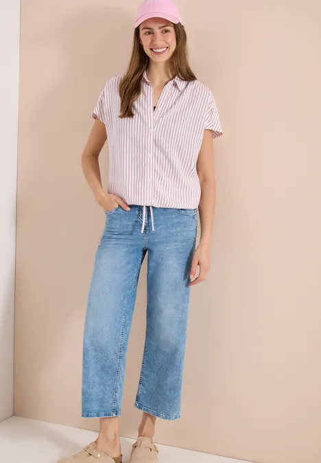 Vrouw in gestreept hemd, wijde jeans en sneakers voor een beige achtergrond.