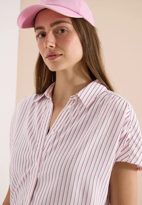 Een persoon draagt een roze pet en een witte blouse met roze strepen voor een beige achtergrond.