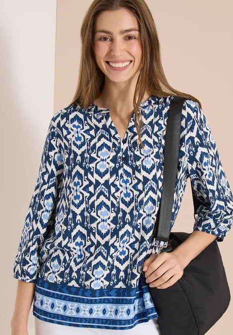 Vrouw in blauw-wit dessinblouse met driekwartmouwen en zwarte schoudertas.
