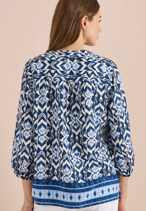 Vrouw van achteren in een losvallende blauw-witte blouse met patroon en driekwart mouwen op neutrale achtergrond.