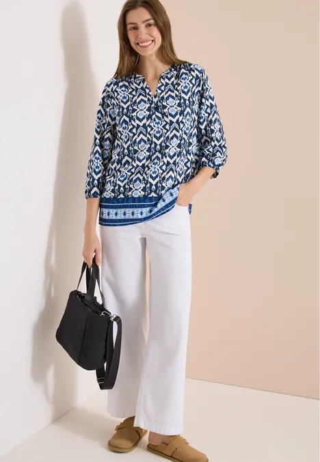 Vrouw met blauw-witte gemêleerde blouse, witte wijde broek, beige loafers en zwarte tas tegen neutrale achtergrond.