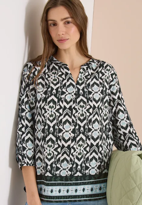 Een vrouw draagt een zwart-wit blouse met geometrische print, driekwart mouwen en een groene zoom.