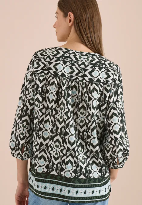 Vrouw van achteren in zwart-wit blouse met geometrisch motief, driekwartsmouwen en groene bies.
