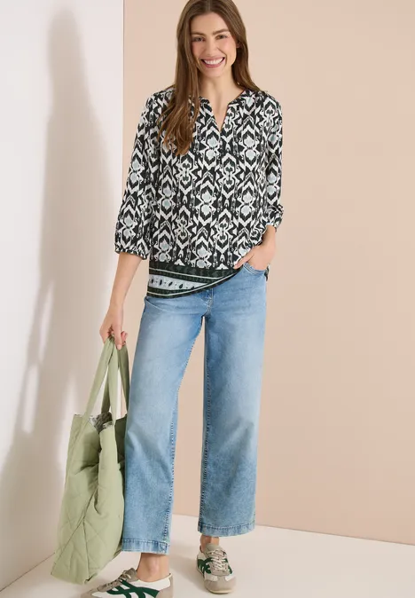 Vrouw met zwart-wit gebloemde blouse, lichtblauwe jeans, sneakers en groene stoffen tas.