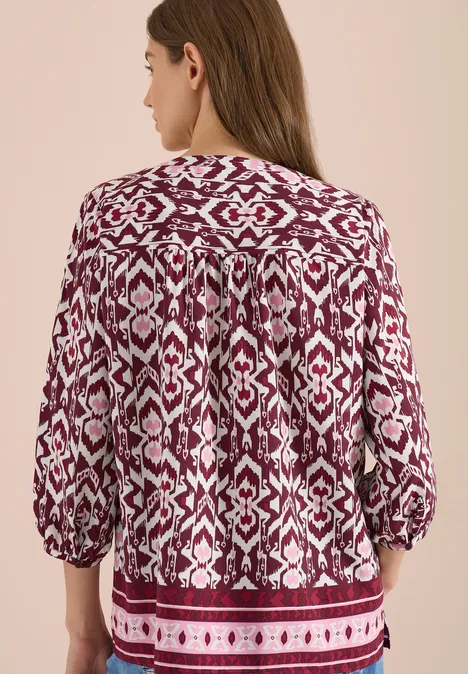 Achteraanzicht van een vrouw in een los vallende rood-wit getekende blouse met driekwartmouwen, beige achtergrond.