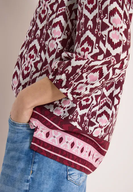 Rode blouse met etnisch patroon, driekwartmouw, hand in de spijkerbroekzak.