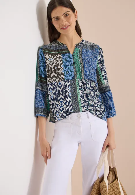 Vrouw in blauw-groene patchworkblouse met witte broek en tas.