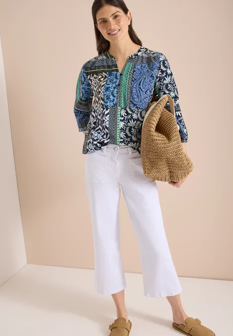 Vrouw met blauwe gebloemde blouse, witte wijde jeans, strooien tas en bruine sandalen.