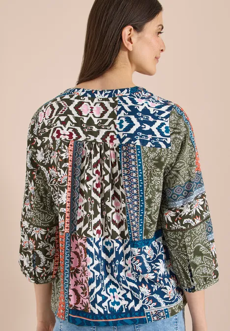 Vrouw draagt een kleurrijk patchwork-blouse met bloemen- en geometrische patronen en 3/4-mouwen.