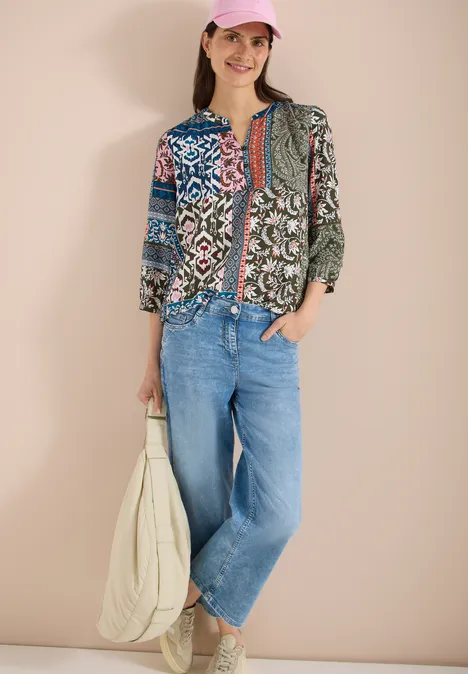 Vrouw voor beige achtergrond draagt kleurrijke patchworkblouse, wijde spijkerbroek en witte tas.