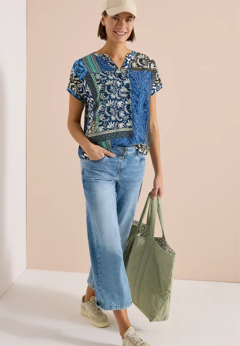 Une femme porte un chemisier bleu à motifs, un jean court clair, des baskets beiges et un grand cabas vert.