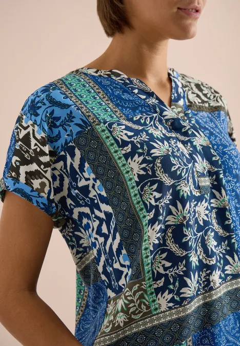 Femme portant un chemisier à manches courtes patchwork bleu et vert sur fond neutre.