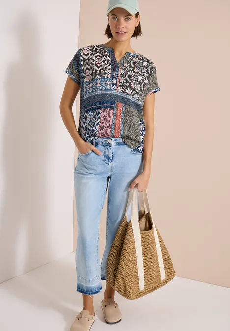 La femme porte un T-shirt imprimé, un jean bleu clair, un sac paille et des espadrilles.