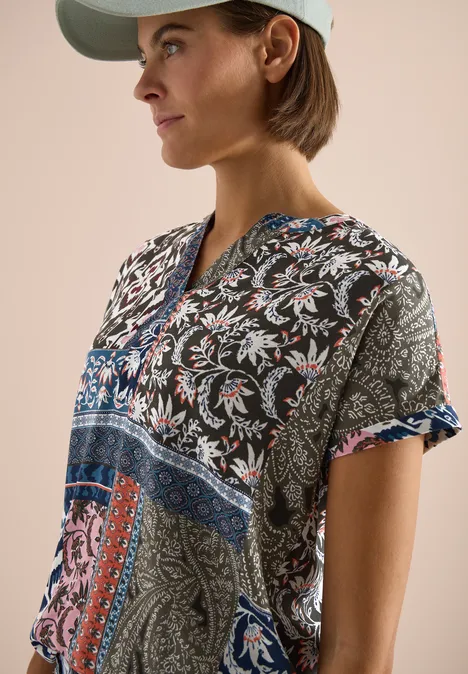 Personne avec casquette portant une tunique colorée à motifs paisley et col en V sur fond beige.