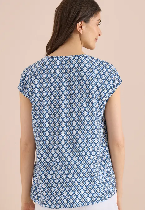 Vrouw van achteren in een blauw-wit geometrisch topje met korte mouwen tegen een lichte achtergrond.