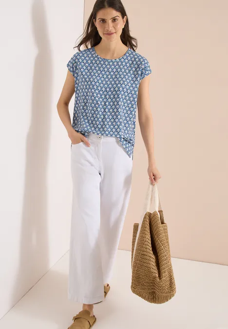 Vrouw draagt blauw top met print, witte wijde broek en grote strooien tas.