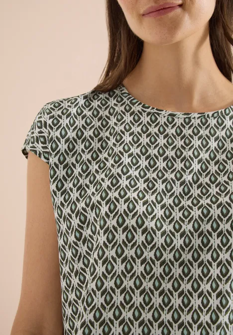 Une femme porte un haut vert à manches courtes avec un motif losange blanc et vert sur fond beige.