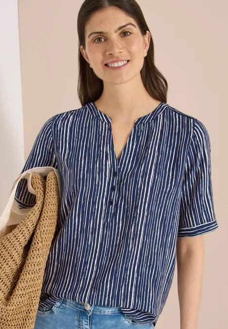 Vrouw in blauw-wit gestreept hemd en witte broek met een gevlochten tas voor een lichte achtergrond.