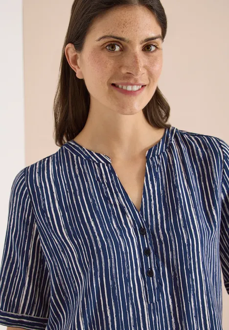 Vrouw draagt blauw-wit gestreept blouse, korte mouwen en knoopsluiting, voor een neutrale achtergrond.