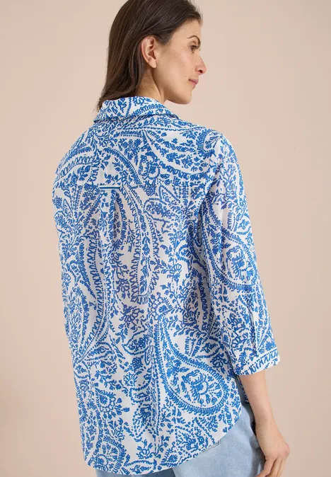 Vrouw van achter gefotografeerd in een blauw-wit paisleypatroon blouse met driekwartmouwen voor een neutrale achtergrond.