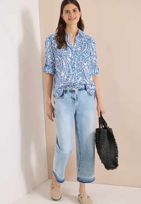 Vrouw leunt tegen muur, draagt blauw patroonblouse, losse jeans, comfortabele schoenen en zwarte gevlochten tas.