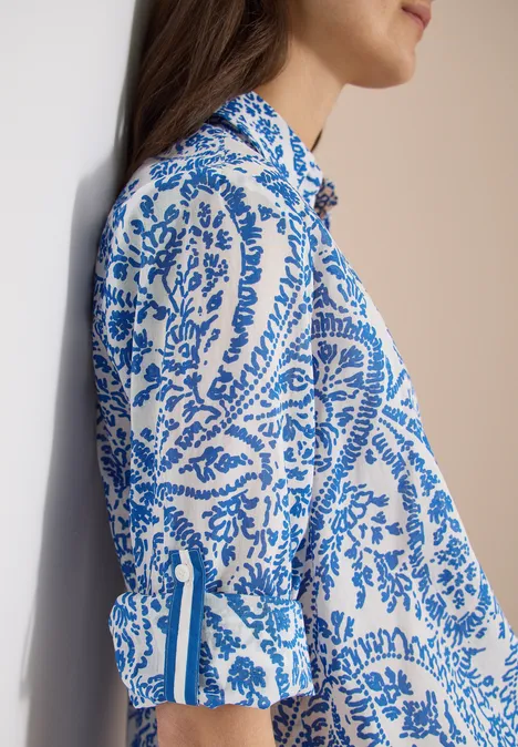 Zijaanzicht van een vrouw in een witte blouse met blauw paisley-patroon, leunend tegen een muur.