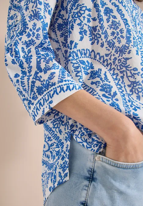 Vrouw in witte blouse met blauw paisley-patroon, hand in de zak van haar lichtblauwe jeans.