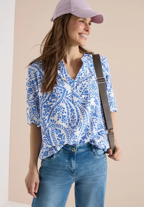 Frau mit abgeflachter Mütze trägt weiße Bluse mit blauem Paisley-Muster, halblangen Ärmeln, Jeans und Umhängetasche.