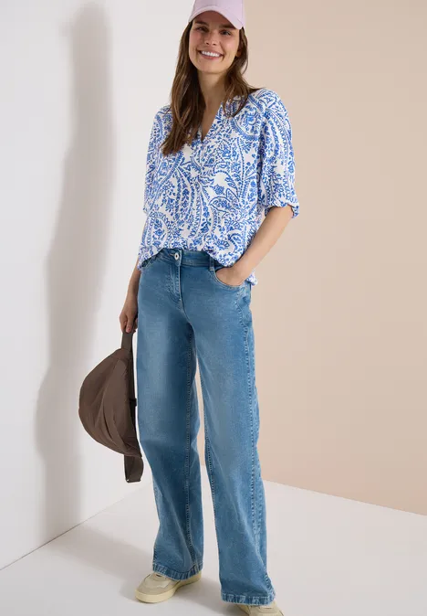 Frau trägt blaue Paisley-Bluse und weite Jeans, hält lässig einen braunen Hut.