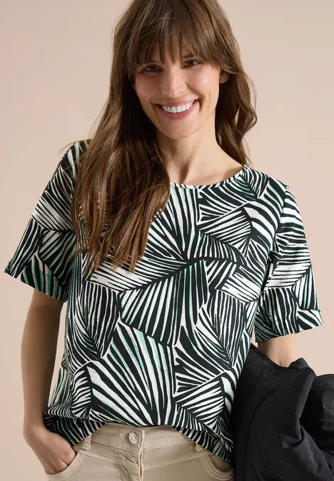Eine Frau trägt ein kurzärmeliges Shirt mit schwarz-weißem Palmenmuster und hält eine Jacke.