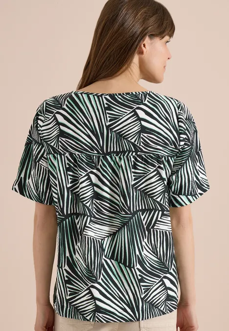 Frau von hinten in kurzärmeliger Bluse mit grün-weiß-schwarzem Blattmuster.