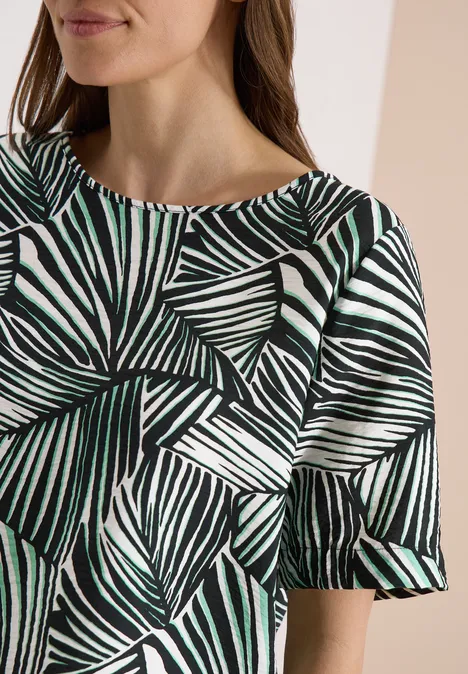Frau in Top mit grünem und schwarzem Blattmuster und kurzen, leicht ausgestellten Ärmeln.