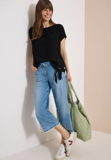 Blouse avec col rond et détail de nœuds Black Femme en t-shirt noir, jean large court, baskets blanches à détail rouge et sac vert en tissu devant un mur clair.