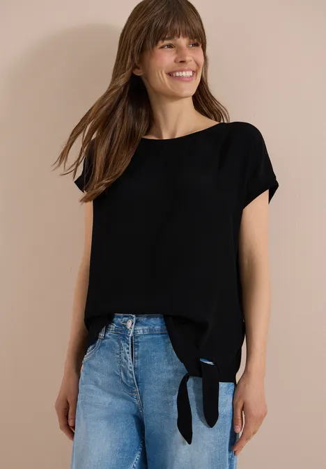 Blouse avec col rond et détail de nœuds Black Une femme porte un haut noir à manches courtes avec un nœud latéral et un jean bleu.