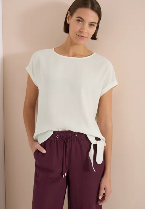 Person porte un top blanc à manches courtes avec nœud sur le côté et pantalon bordeaux.