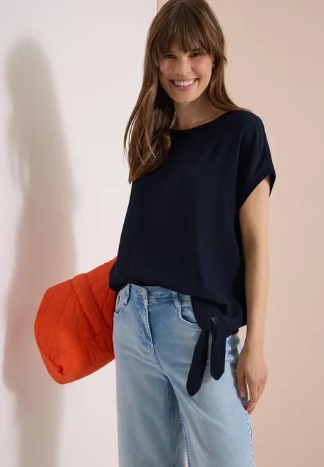 Blouse avec col rond et détail de nœuds universal blue Femme en t-shirt bleu marine noué sur le côté, jean clair, portant un grand sac rouge décontracté.