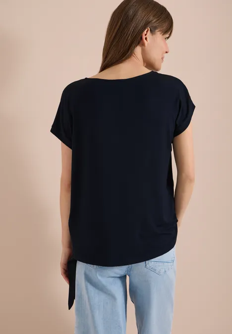 Blouse avec col rond et détail de nœuds universal blue Femme de dos portant un t-shirt bleu foncé et un jean bleu clair sur fond neutre.