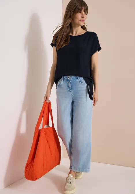 Blouse avec col rond et détail de nœuds universal blue Femme appuyée contre un mur, porte un top noir, jean bleu clair, baskets blanches et un grand sac rouge.