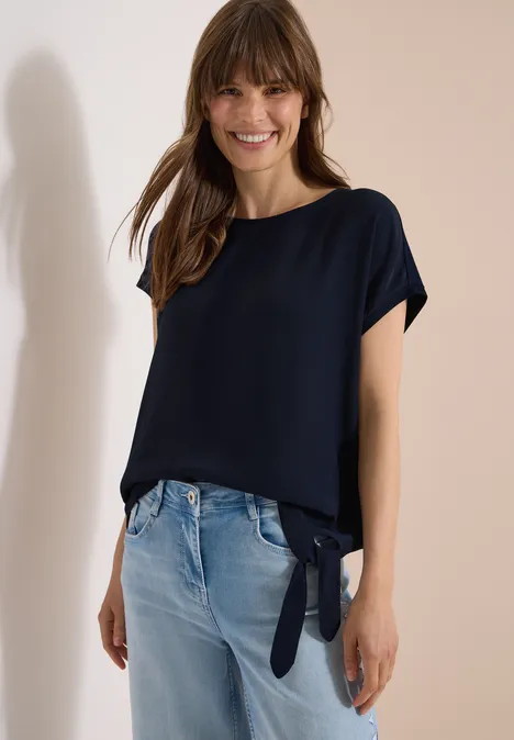 Blouse avec col rond et détail de nœuds universal blue Femme portant un top bleu marine ample noué sur le côté et un jean clair, posant devant un fond clair.