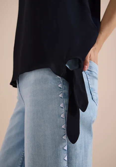 Blouse avec col rond et détail de nœuds universal blue Top noir et jean clair avec incrustations triangulaires sur le côté, main dans la poche.