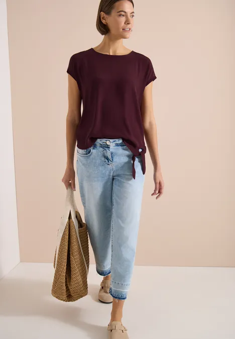 Femme en t-shirt bordeaux et jean clair, tenant un sac en paille, sur fond neutre.