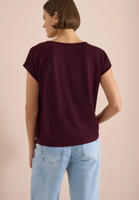 Vue de dos d’une femme portant un t-shirt bordeaux et un jean bleu clair sur fond neutre.
