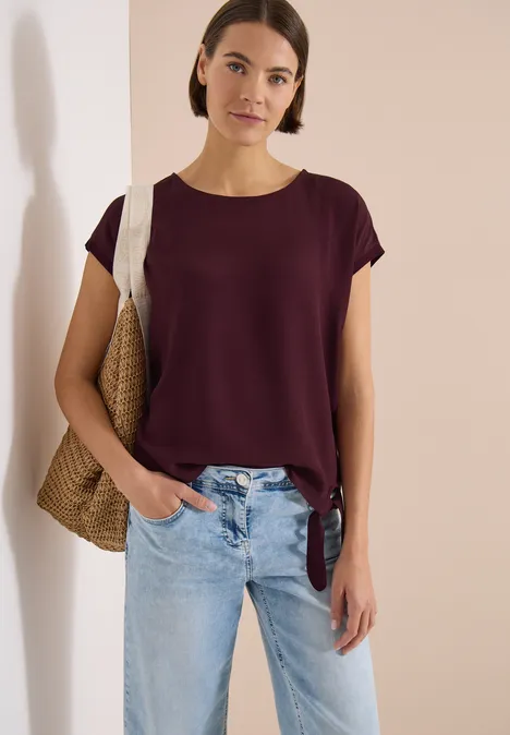 Personne en T-shirt violet, jean bleu et sac en paille, debout de manière décontractée devant un mur blanc.