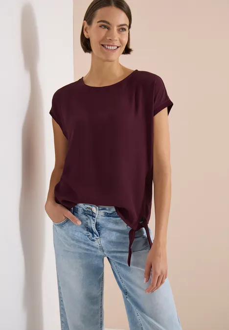 Une femme porte un t-shirt bordeaux noué sur le côté et un jean clair.