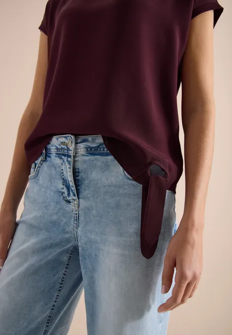 Femme porte un chemisier bordeaux, noué sur le côté, avec un jean bleu clair ample.