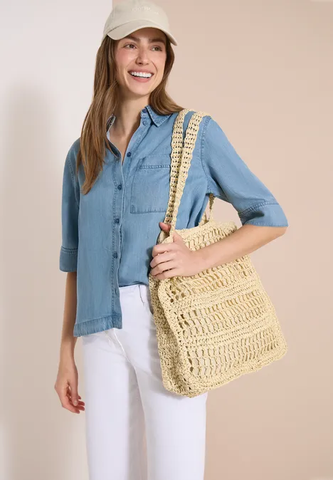 Vrouw met lichtblauw overhemd, witte broek en grote gevlochten strooien tas.