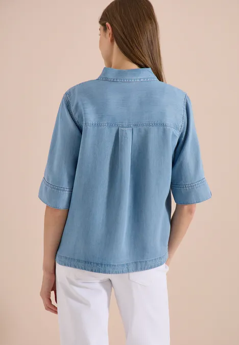 Achteraanzicht van een vrouw in een lichtblauwe spijkerblouse en witte broek.