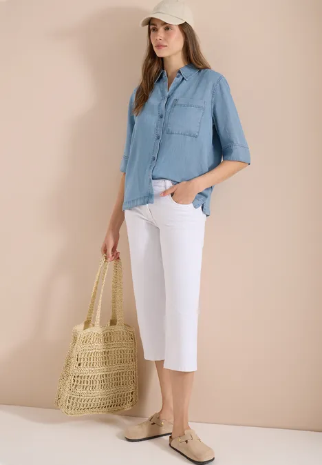 Vrouw draagt lichtblauw overhemd, witte culottes, beige ballerina's en houdt een gevlochten strooien tas.