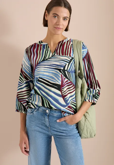 Frau mit bunter Bluse im Blattmuster, blauen Jeans und hellgrüner gesteppter Tasche vor beigem Hintergrund.